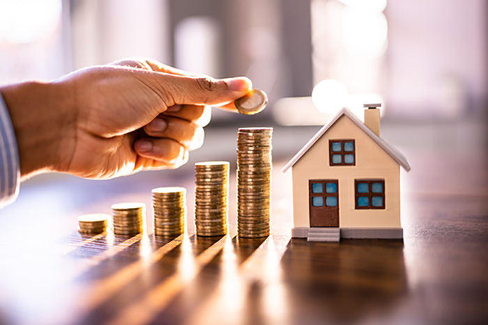 Hand placing a coin on a rising stack of coins beside a small house model, representing real estate investment, savings, or property financing growth.