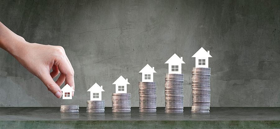 Hand placing a small house icon on a stack of coins, with additional house icons on rising coin stacks to show real estate growth.