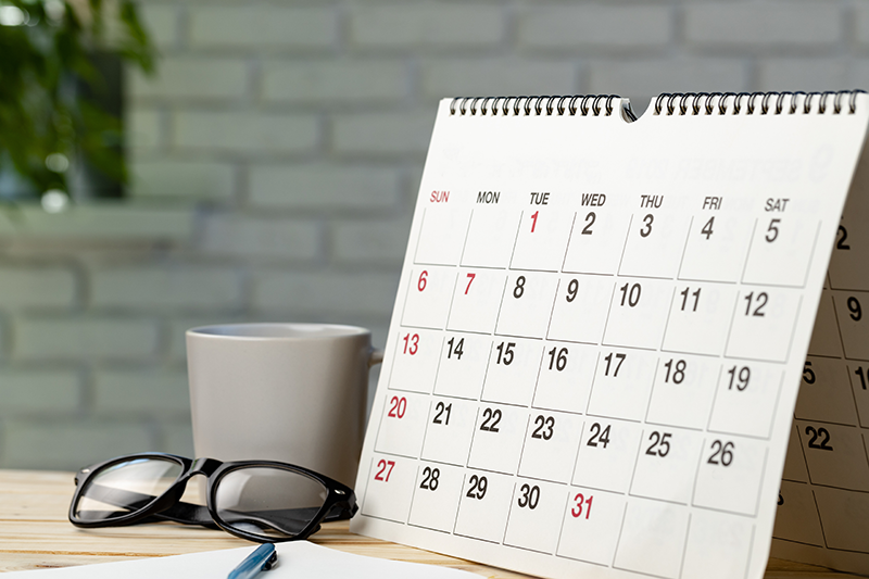 Desk calendar showing a monthly schedule beside a coffee mug, eyeglasses, and a pen on a wooden table.